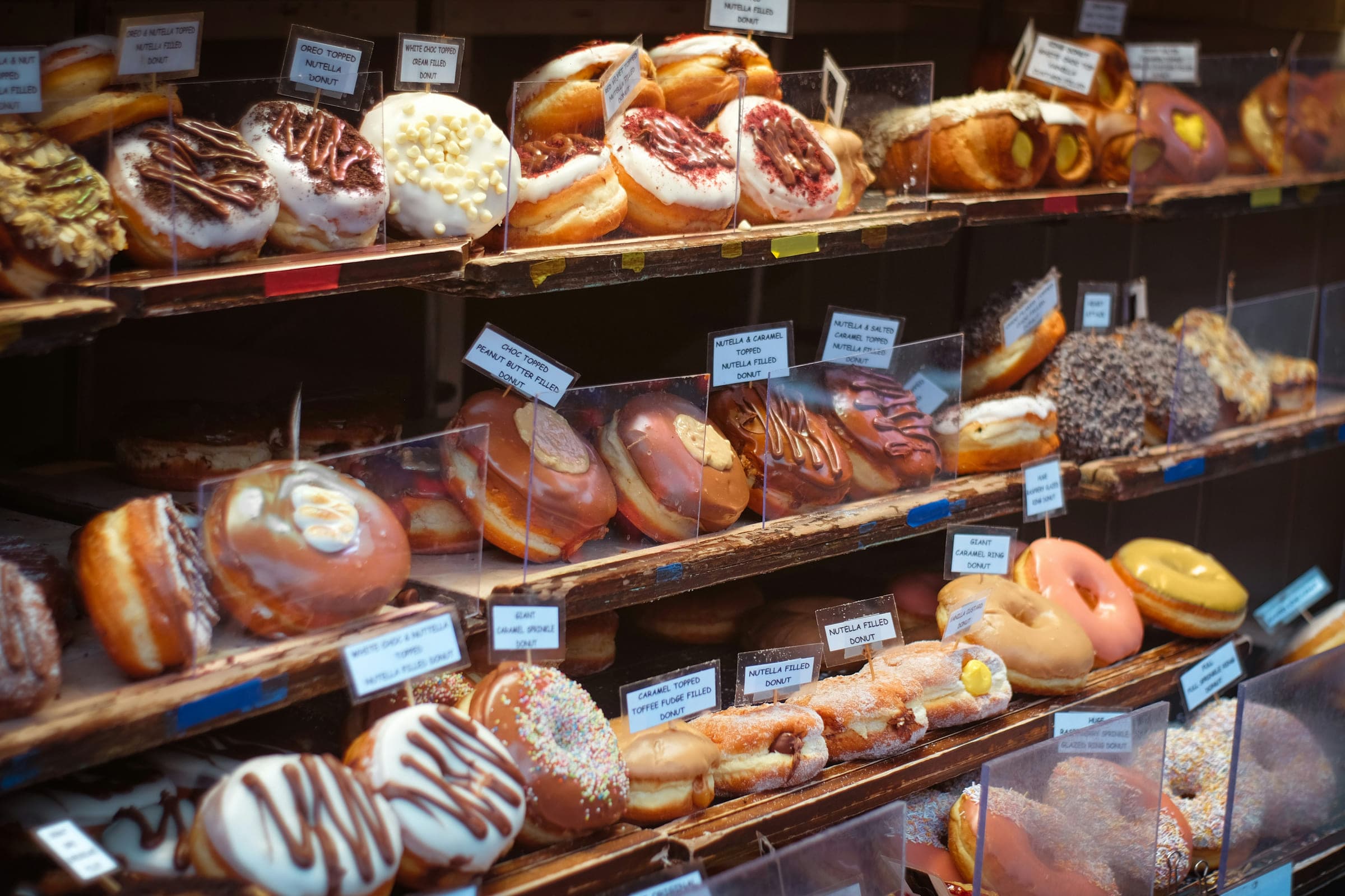 よその店のドーナツのケース。木の棚が三段、ガラスの向こうに何十種類ものドーナツと、品名を書いた札が立っている。
